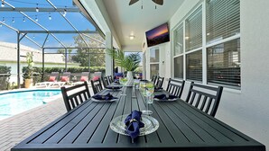 A covered patio with a pool, dining table set with plates and napkins, and a TV mounted on the wall.
