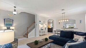 A modern living room with a dark blue sectional sofa, a wooden coffee table, and a blue patterned armchair. A staircase leads to an upper level, and a kitchen area is visible in the background.