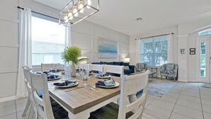 A dining area with a wooden table set for four, white chairs with green cushions, a potted plant, and a large window with blinds.