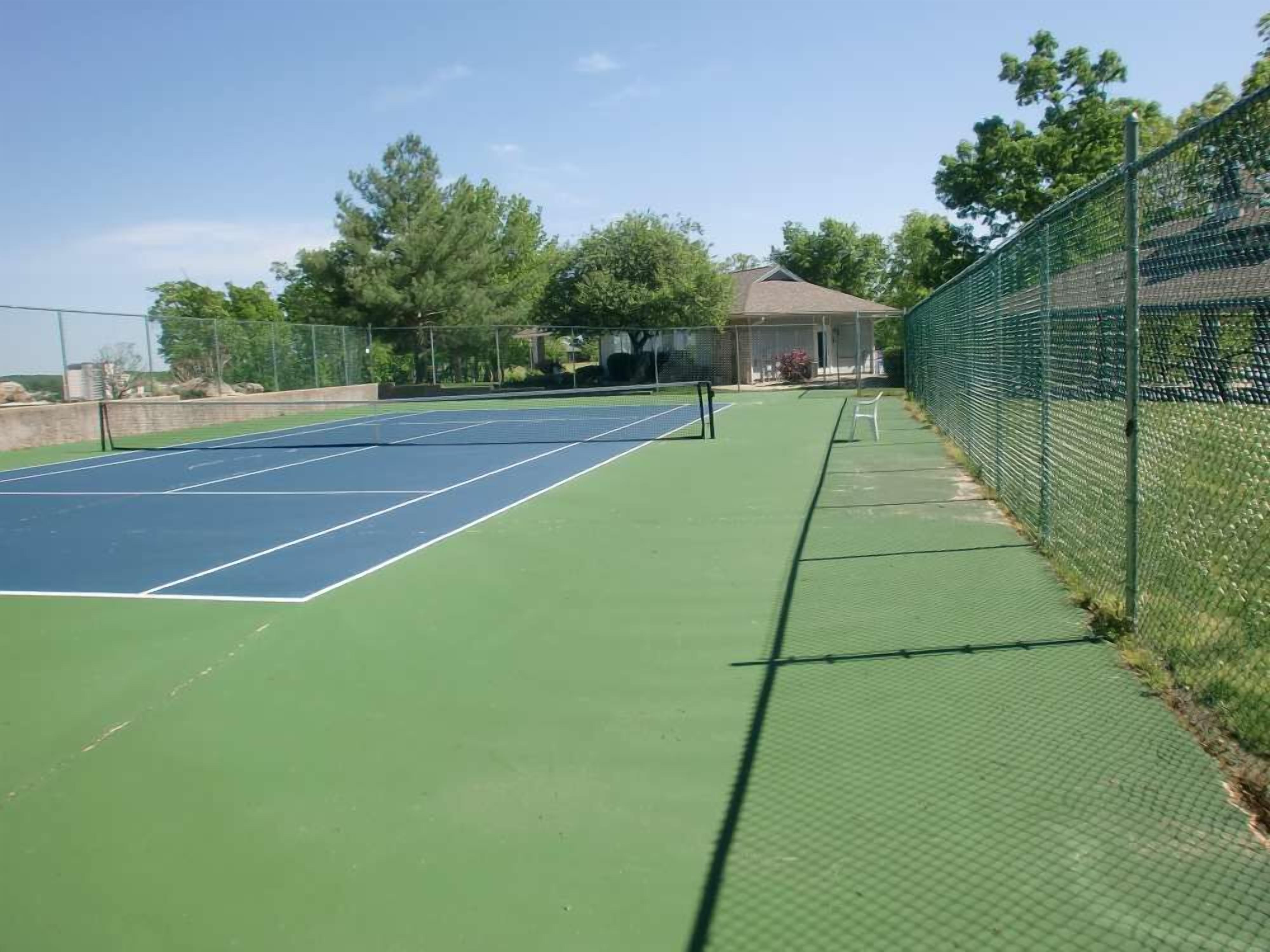 An outdoor tennis court with a green surface, surrounded by a chain-link fence.