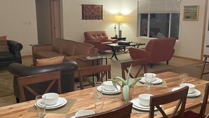 Zona pranzo con tavolo in legno apparecchiato per quattro, circondato da sedie di legno. Sullo sfondo, una poltrona in pelle marrone e una poltrona rossa.