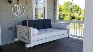 Een veranda met een witte schommelstoel en kussens, een witte deur met een sleutelgat, en een raam met een grillwerk ontwerp.