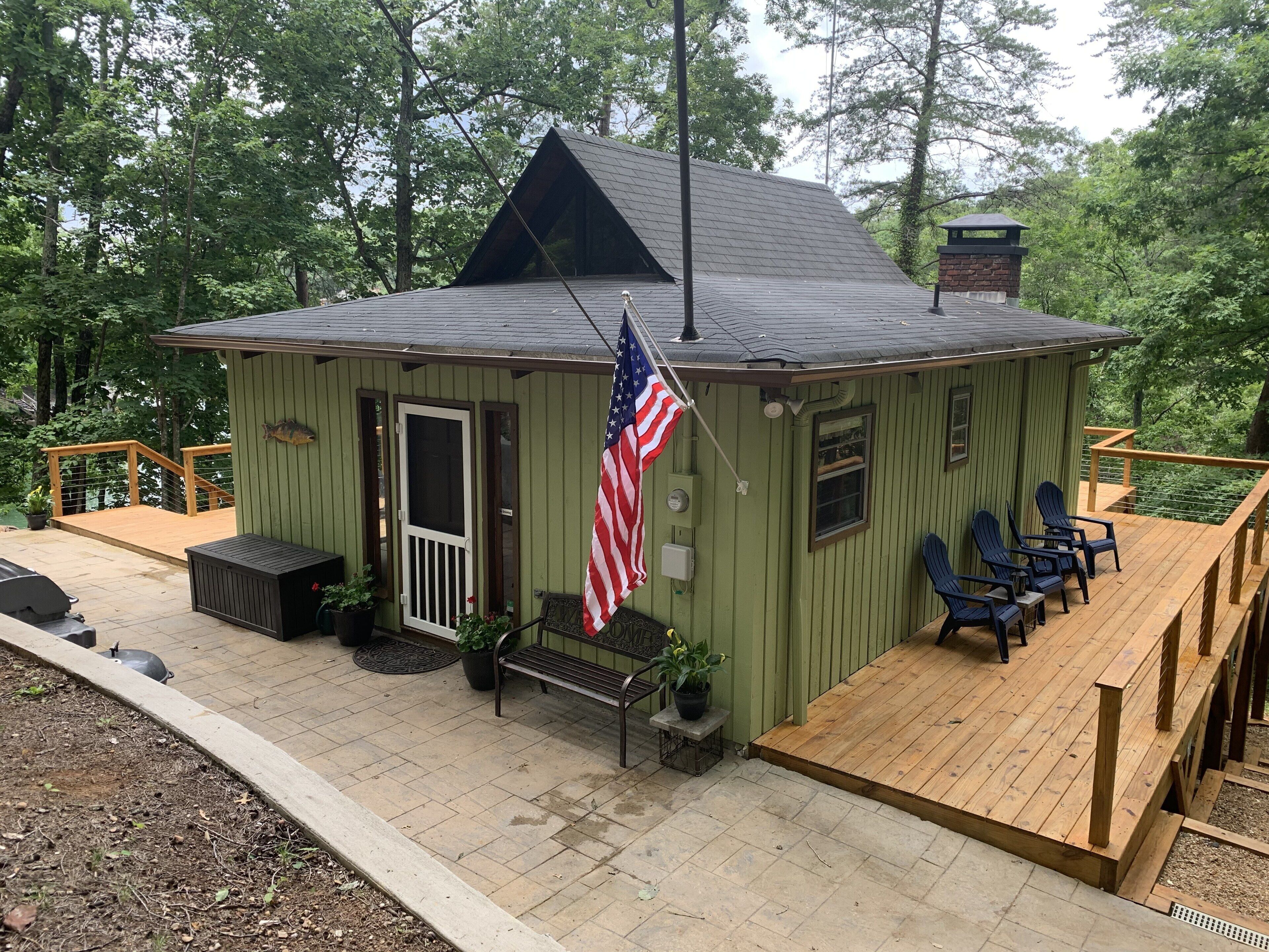 Een kleine, groene blokhut met een houten terras, een Amerikaanse vlag en buitenmeubels.