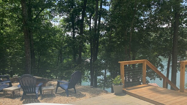 A hot tub surrounded by outdoor furniture, a wooden deck, and a view of a lake with trees.