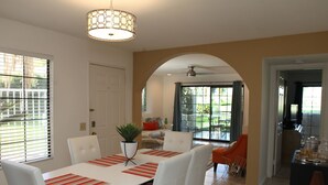 Un coin repas avec une table blanche, des chaises blanches et une plante en pot. Une porte-fenêtre coulissante donne accès à un patio aménagé avec du mobilier d’extérieur.