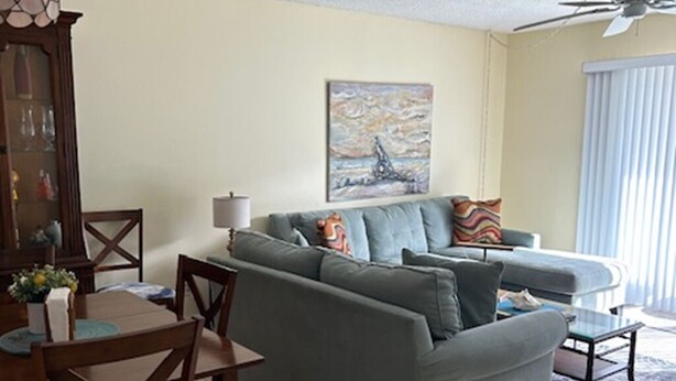 Een woonkamer met een grijze bank, een houten eettafel en stoelen. Er is een plafondventilator en een schilderij aan de muur.