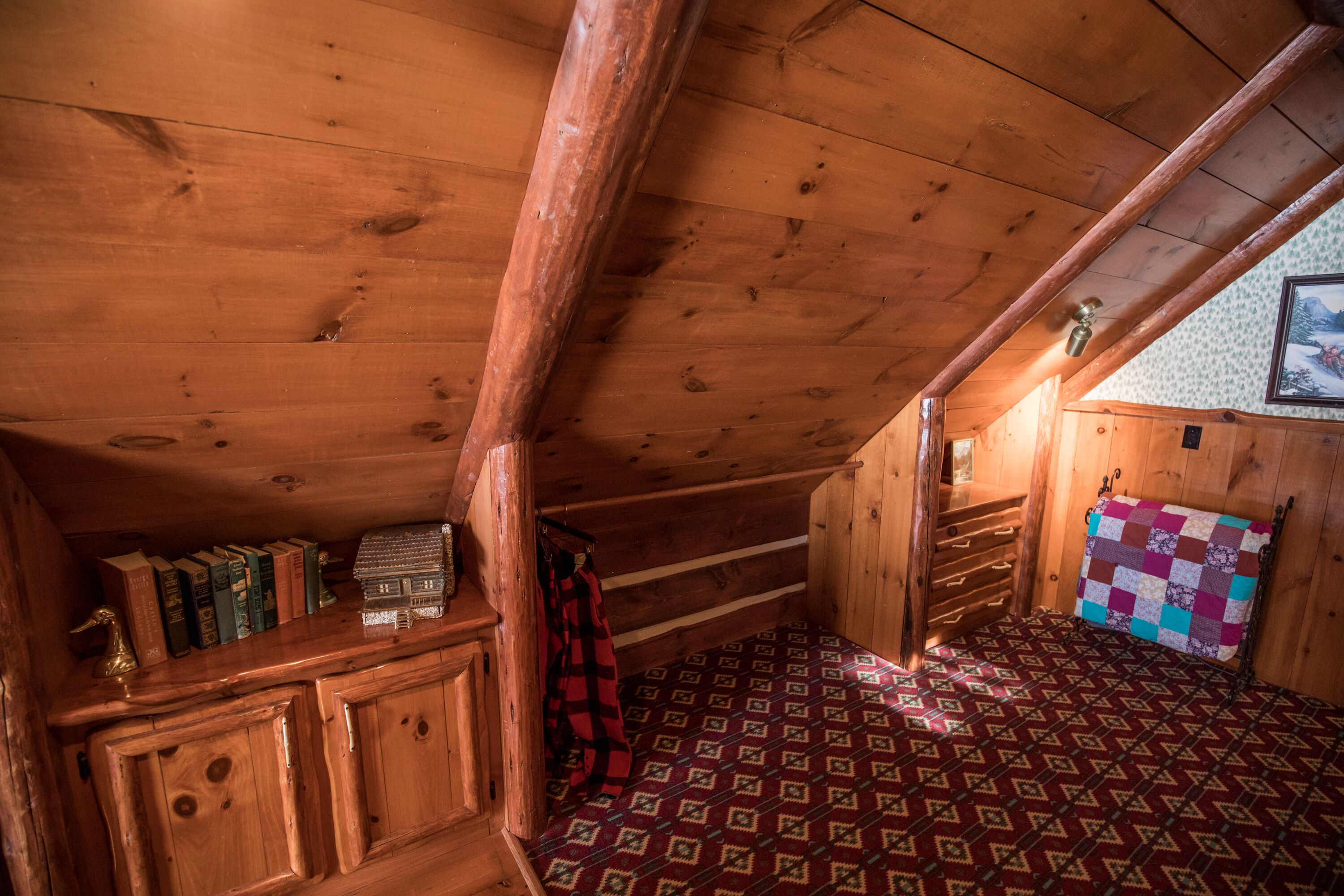 L’intérieur d’un chalet en bois, avec un plafond en pente, un banc en bois, un lit recouvert d’une courtepointe et une bibliothèque contenant des livres et un petit objet décoratif.