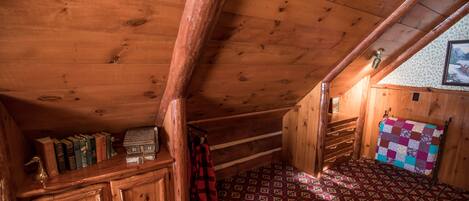 L’intérieur d’un chalet en bois, avec un plafond en pente, un banc en bois, un lit recouvert d’une courtepointe et une bibliothèque contenant des livres et un petit objet décoratif.