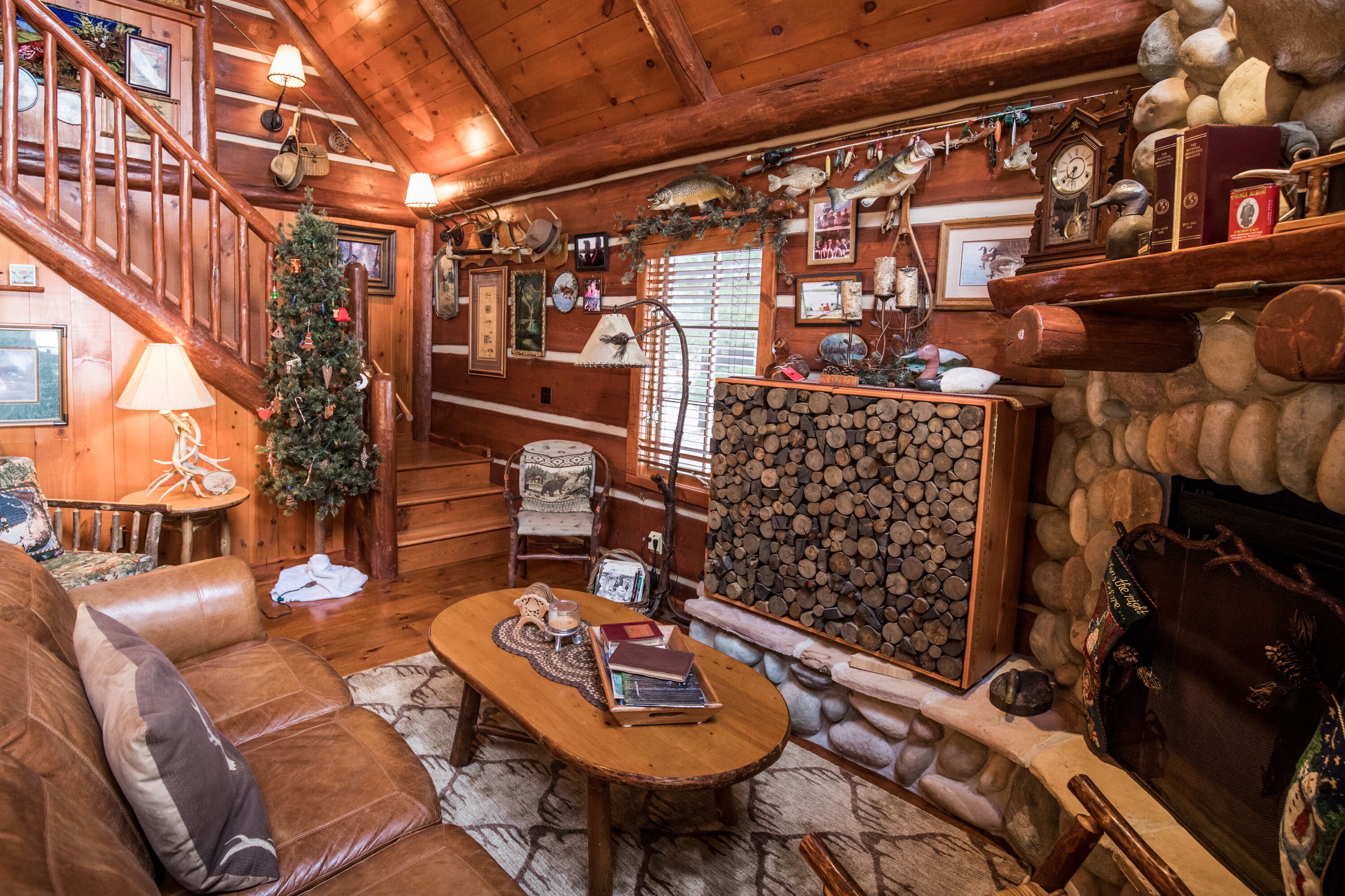 A cozy cabin interior with a wooden staircase, a fireplace, a Christmas tree, and various decorations.