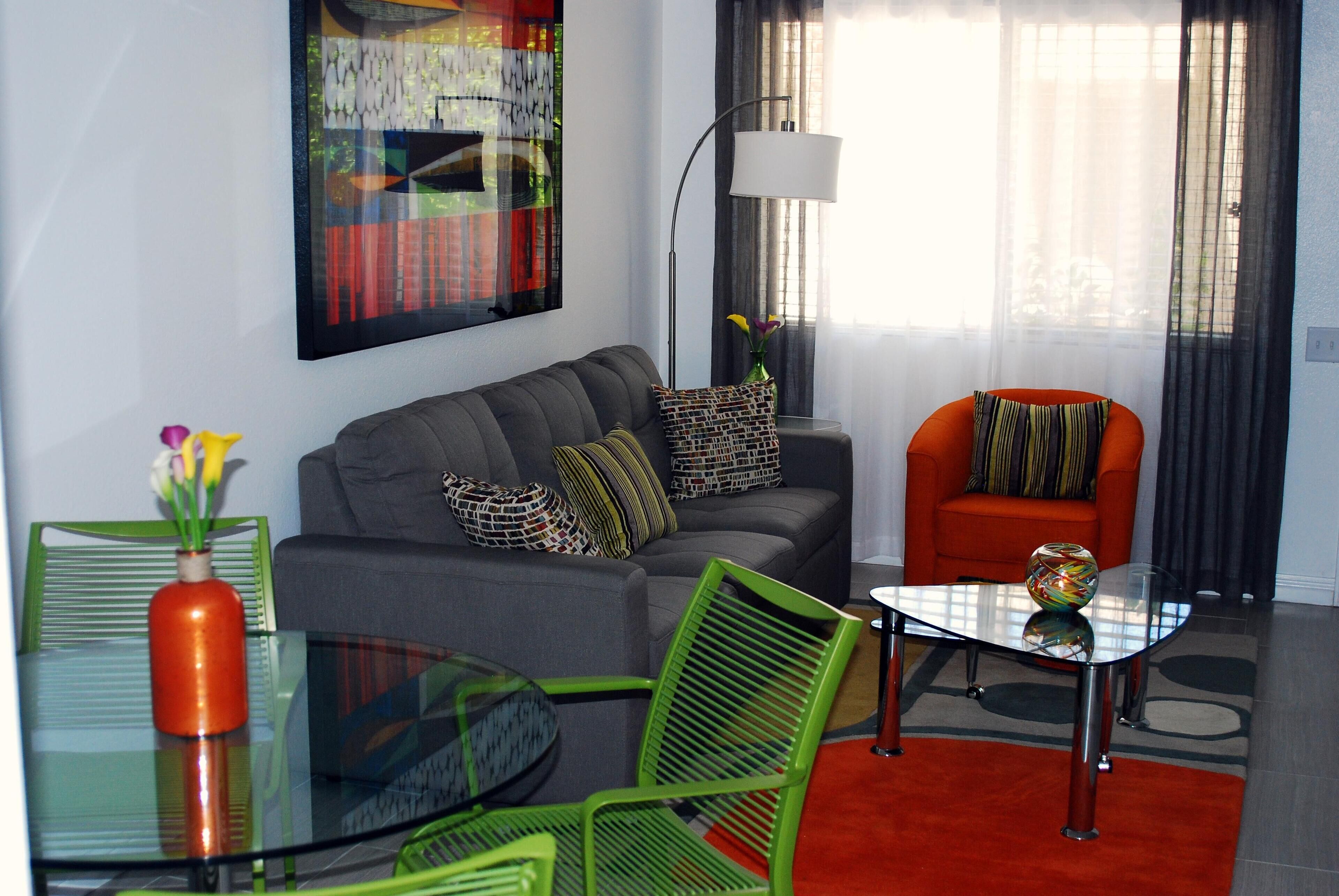 Una sala de estar moderna con un sofá gris, sillas verdes, una mesa de centro de vidrio y un cuadro abstracto colorido en la pared.