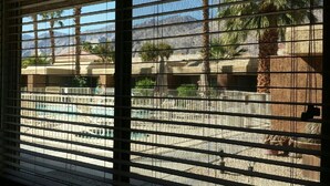 A view through a window with horizontal blinds, featuring a pool area with palm trees and a mountainous landscape in the background.