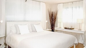 Et hotelværelse med en stor seng, et natbord, et skrivebord og et vindue med lette gardiner.