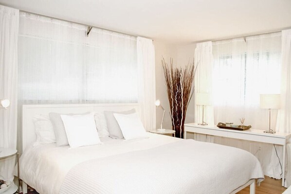 Una habitación de hotel con una cama grande, una mesita de noche, un escritorio y una ventana con cortinas translúcidas.