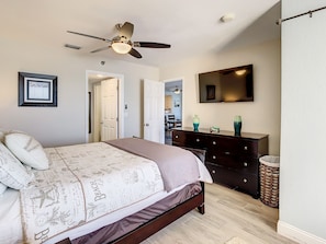 A bedroom with a bed, a dresser, a ceiling fan, a TV, and a door leading to another room.