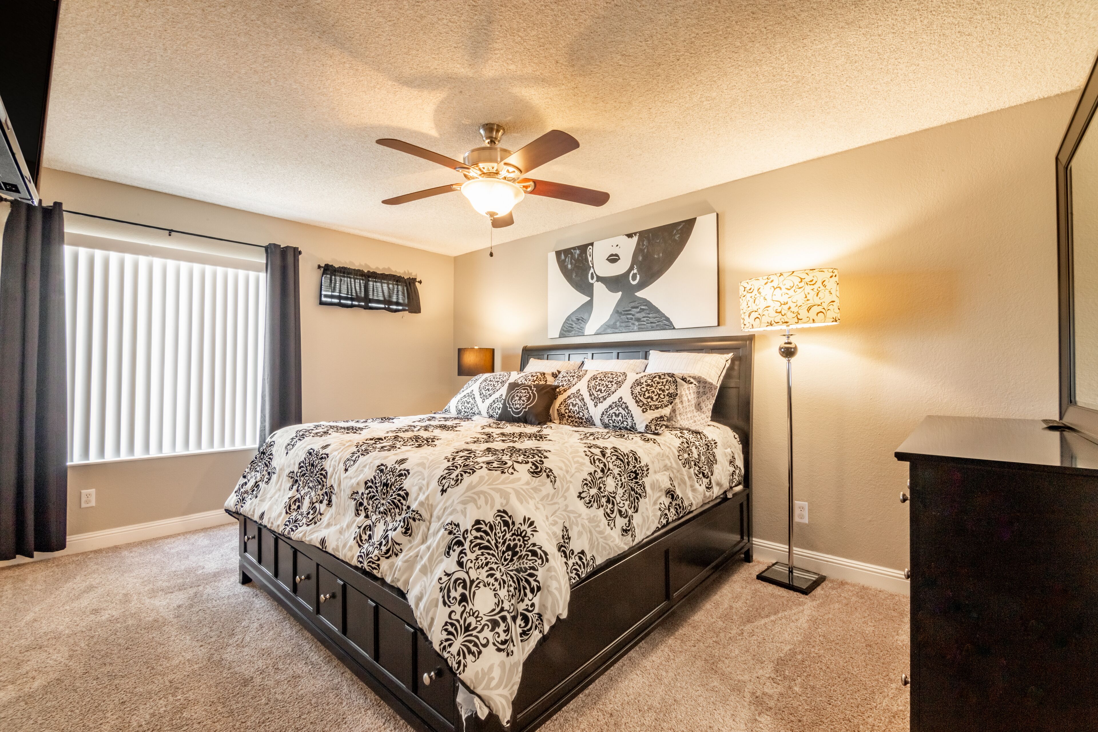 A bedroom with a bed, ceiling fan, artwork, lamp, and a dresser.