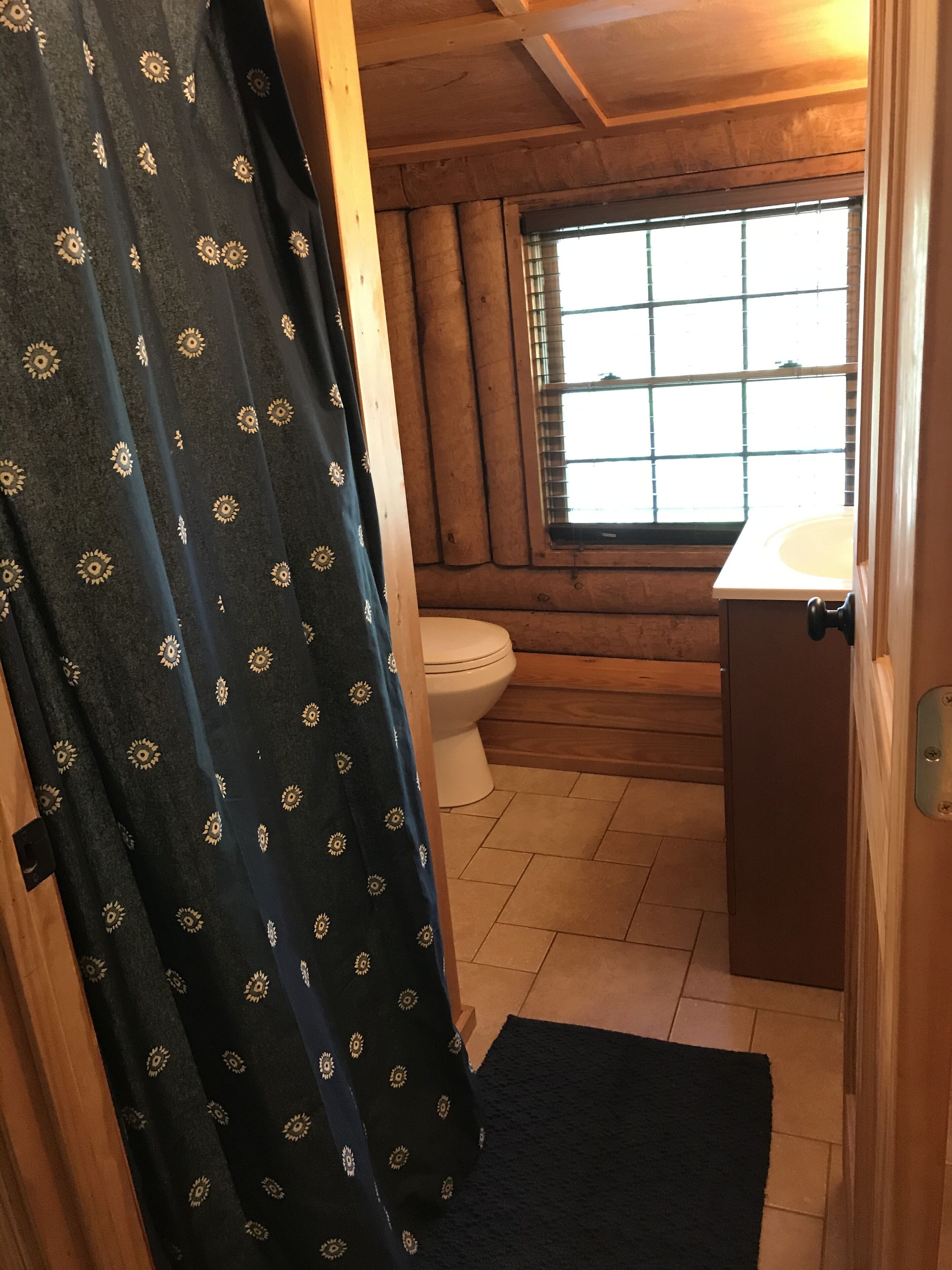 A bathroom with a wooden cabin-style interior, a toilet, a sink, and a window with blinds.
