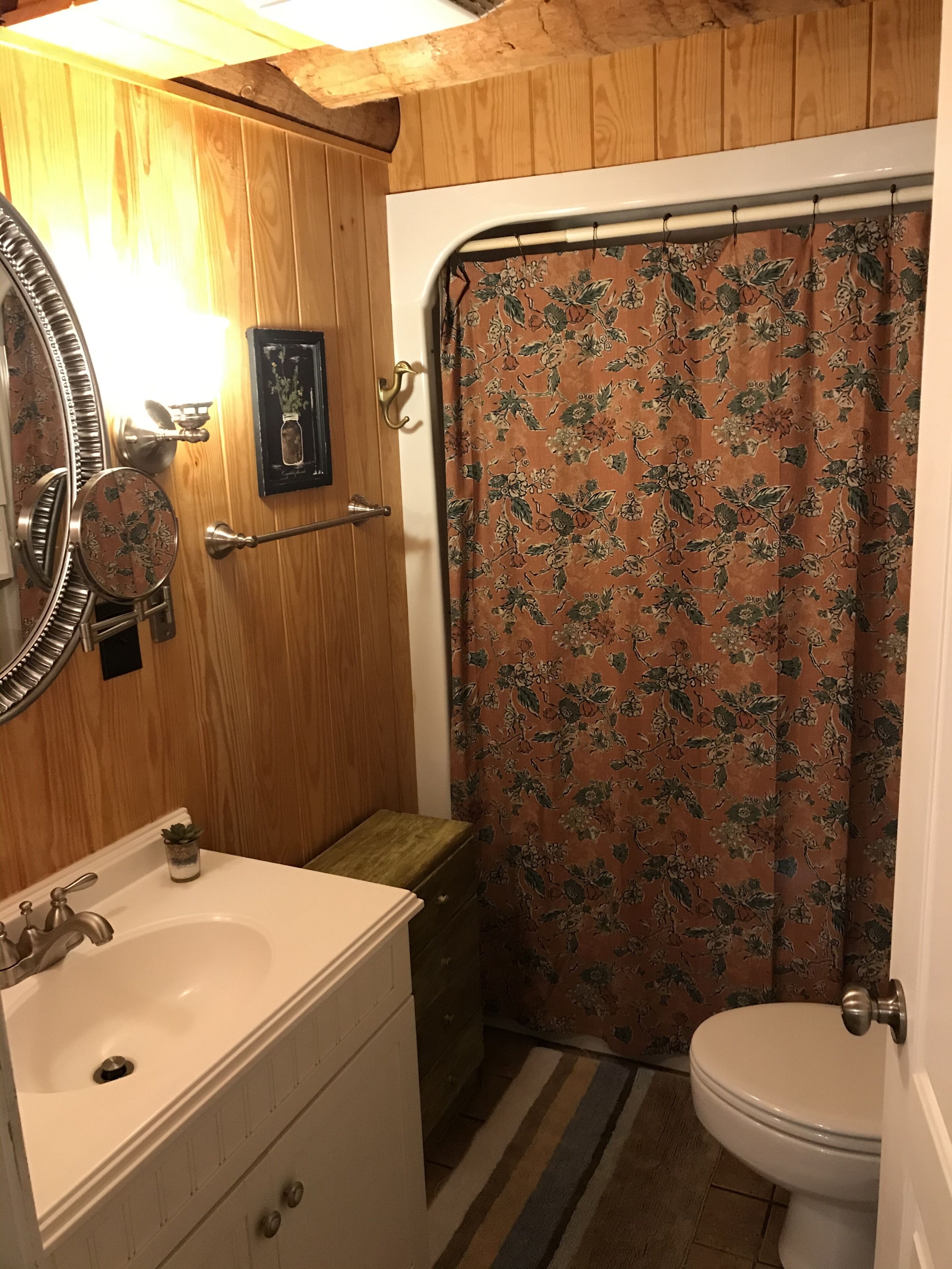Un bagno con un mobiletto vanità in legno, un lavandino, un WC e una tenda da doccia a motivi floreali.