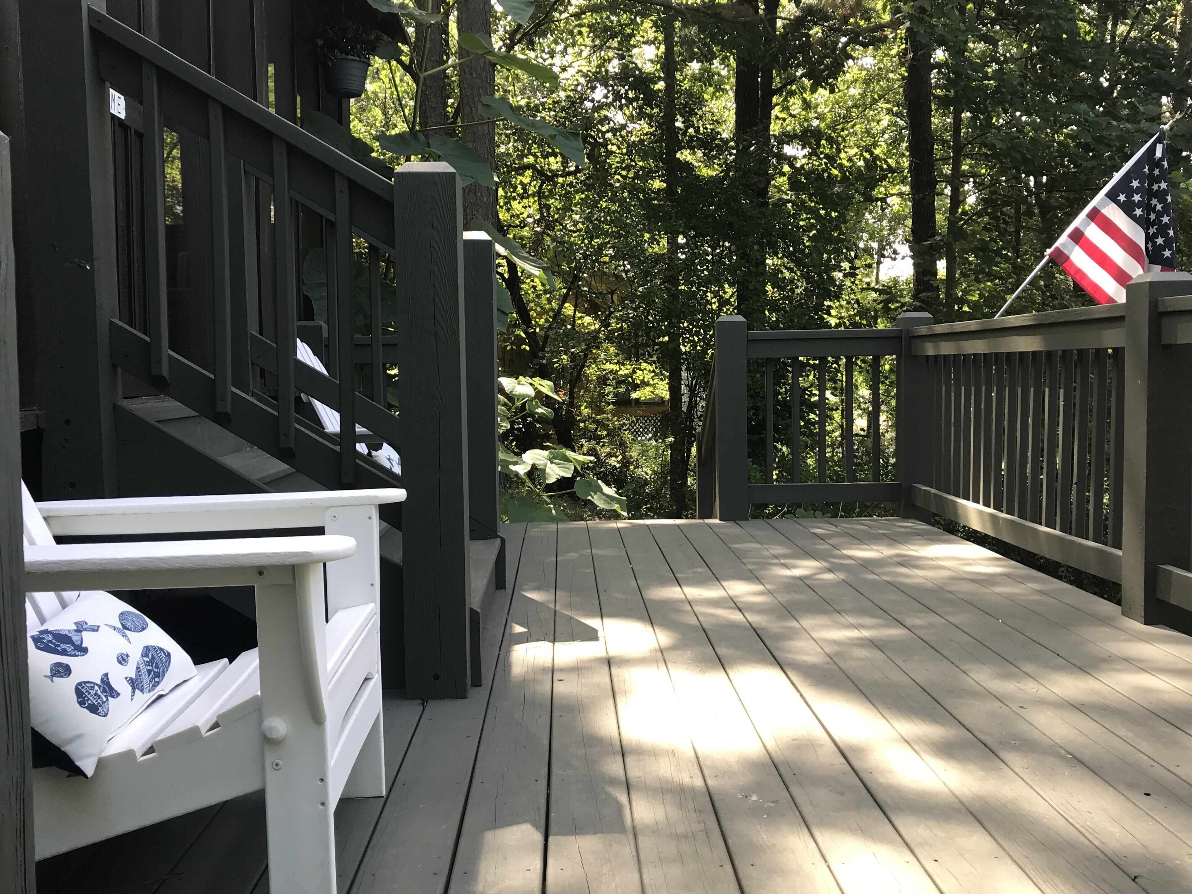 A wooden deck with a white bench and a small table, surrounded by lush greenery and an American flag.