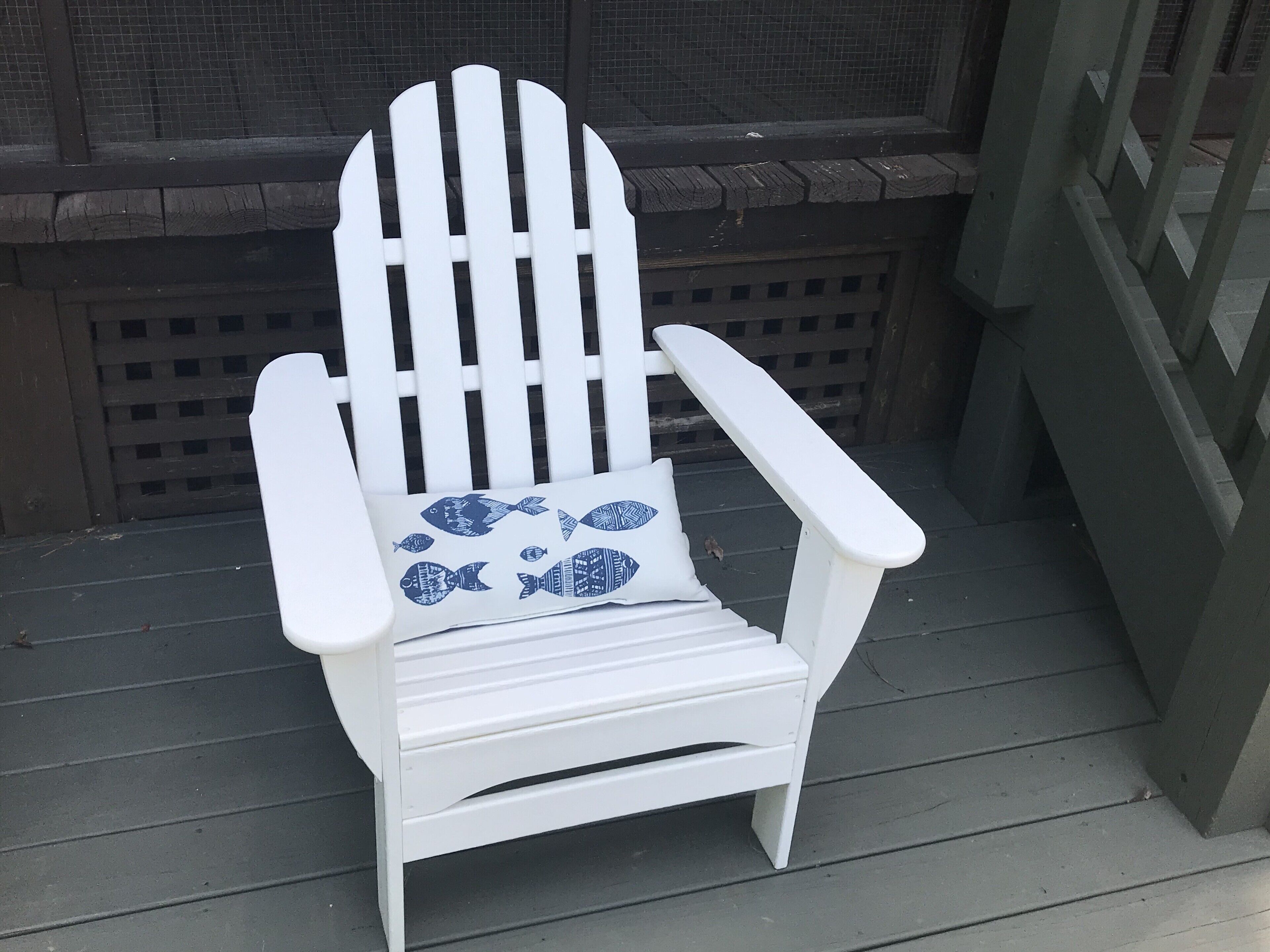 Una cadira Adirondack blanca amb coixí de motius de peixos blaus sobre un pont de fusta.