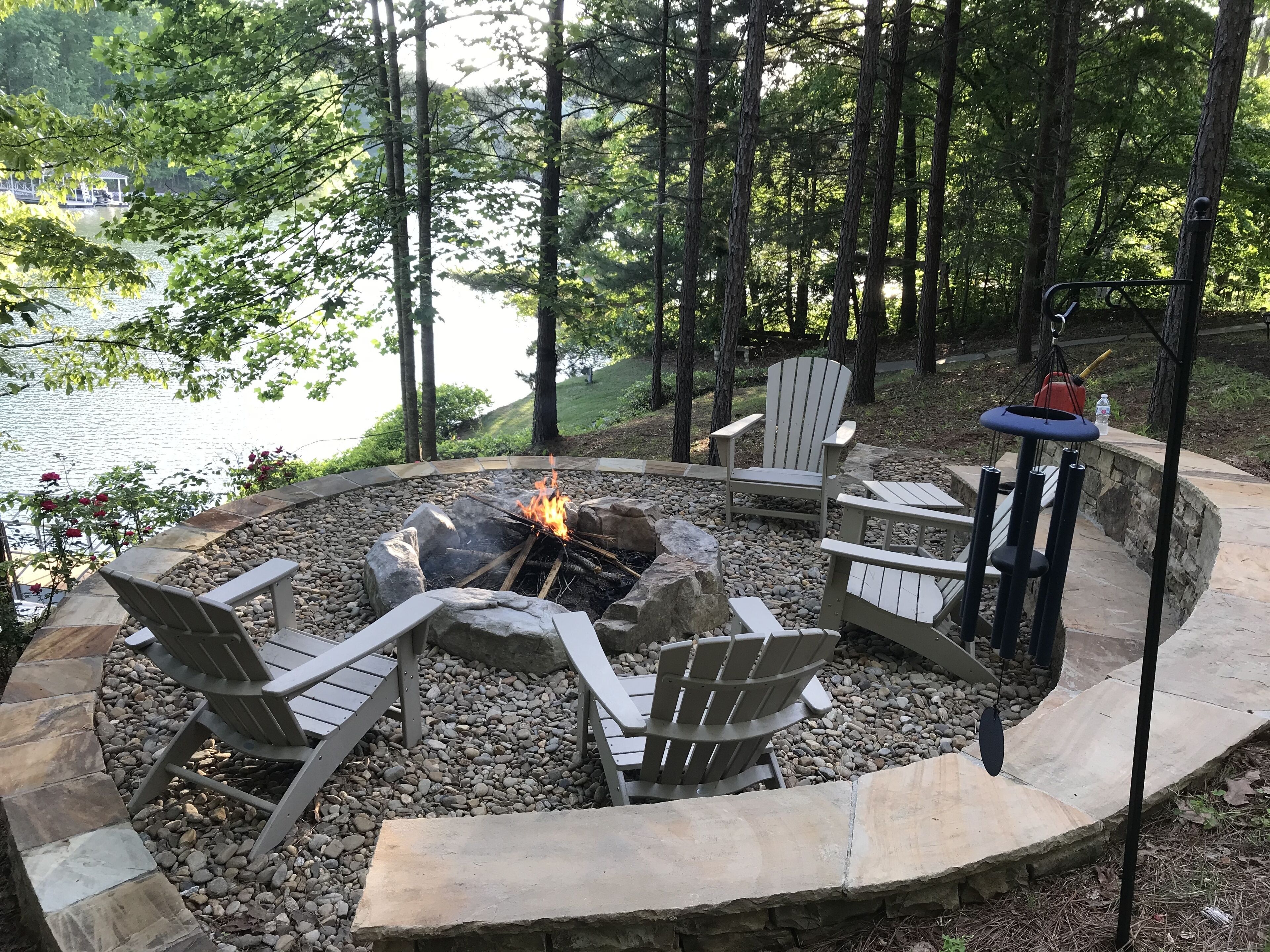 Un espace extérieur circulaire aménagé avec des sièges, doté d’un foyer, entouré de mobilier en pierre et en bois, et offrant une vue sur un lac.