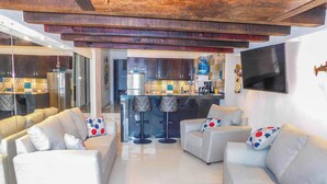 Una sala de estar moderna con sofás blancos, una cocina al fondo y un techo de madera.