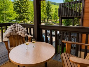 A wooden deck with a round table and two chairs, two glasses of white wine, and a fur throw on one chair.