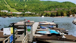 A marina with boats docked, a wooden pier, and a backdrop of green hills and buildings. - RENOVATED- Ski in&out /Walk to village/ Corner Unit/Pool (summer) Sunny/Views/AC (Mont-Tremblant)