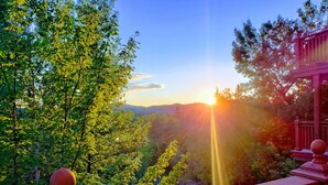 A wooden balcony with a balustrade overlooking a lush valley with trees and a distant view of the sun setting behind hills. - RENOVATED- Ski in&out /Walk to village/ Corner Unit/Pool (summer) Sunny/Views/AC (Mont-Tremblant)