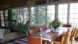 A dining area with a wooden table and wicker chairs, large windows with a view of trees, and a sign indicating no dogs allowed on the beach. - Acadia National Park-Oceanfront Cottage-Views of Cadillac Mountain & Acadia-Pets (Lamoine)
