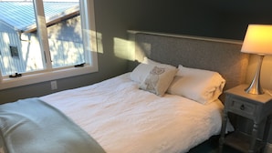 Een net opgemaakt bed met een witte deken en kussens, een bedlampje en uitzicht buiten door een raam.