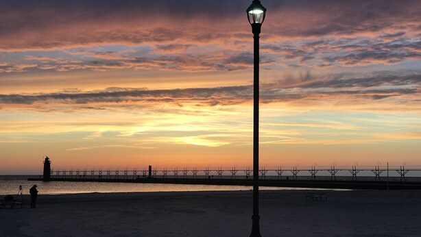 A coastal scene with a lighthouse, a pier, and a beach at sunset. - Only 2 blocks to beach, one block to kids corner, over half an acre! (South Haven)