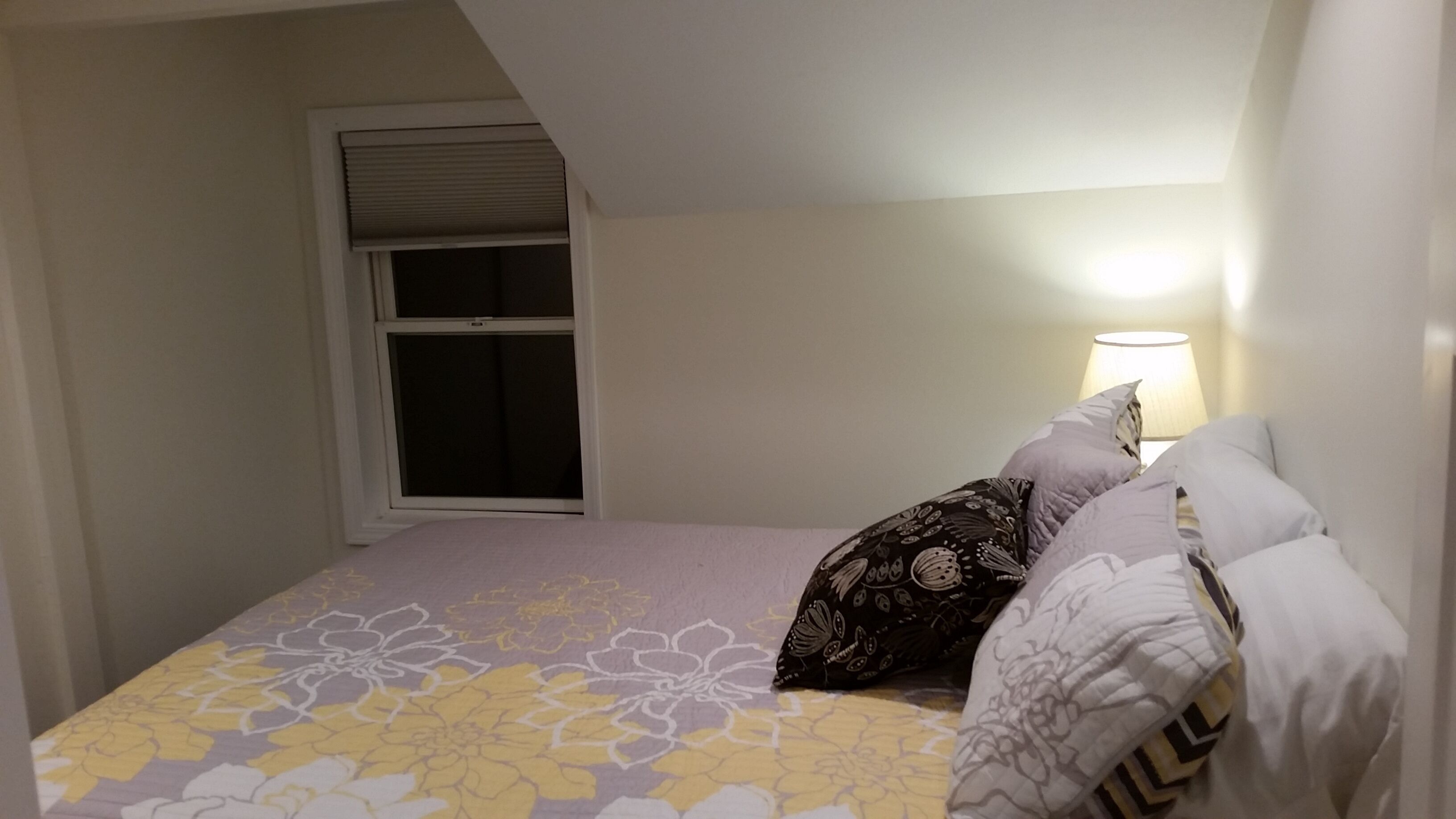 Un dormitorio con cama, almohadas, ventana y lámpara.
