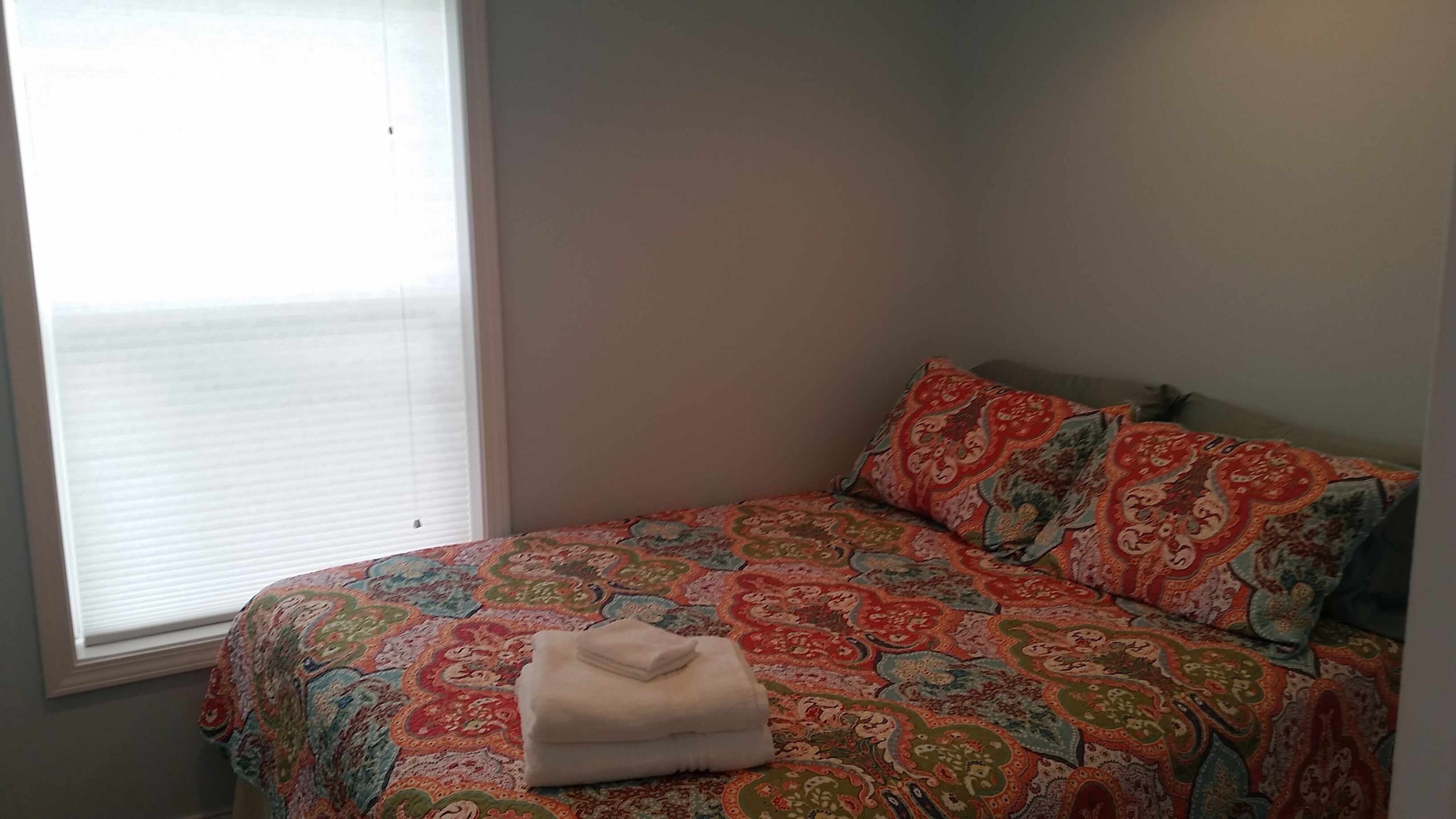Ein Schlafzimmer mit einem Bett, das mit einer gemusterten Tagesdecke bezogen ist, einem Fenster mit Jalousien und einem auf dem Bett liegenden gefalteten Handtuch.