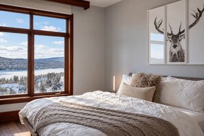 A bedroom with a large window, a bed with white bedding, a fluffy pillow, and a framed deer picture on the wall.