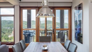 Uma sala de jantar com uma grande mesa de madeira, quatro cadeiras, um lustre pendente e amplas janelas que oferecem vista para uma paisagem montanhosa.