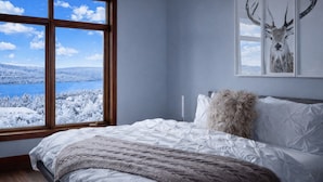 Quarto com uma janela grande que oferece vista para uma paisagem nevada, cama com roupa de cama branca e travesseiro fofo, e uma arte na parede com chifres de veado emoldurados.