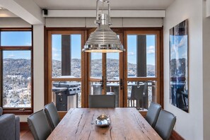 A dining area with a wooden table, chairs, a hanging light fixture, and large windows offering a view of a mountainous landscape.