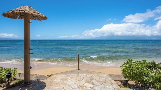 Une plage avec une vue sur l’océan, un parasol en chaume, un sentier pavé et un ciel bleu dégagé.