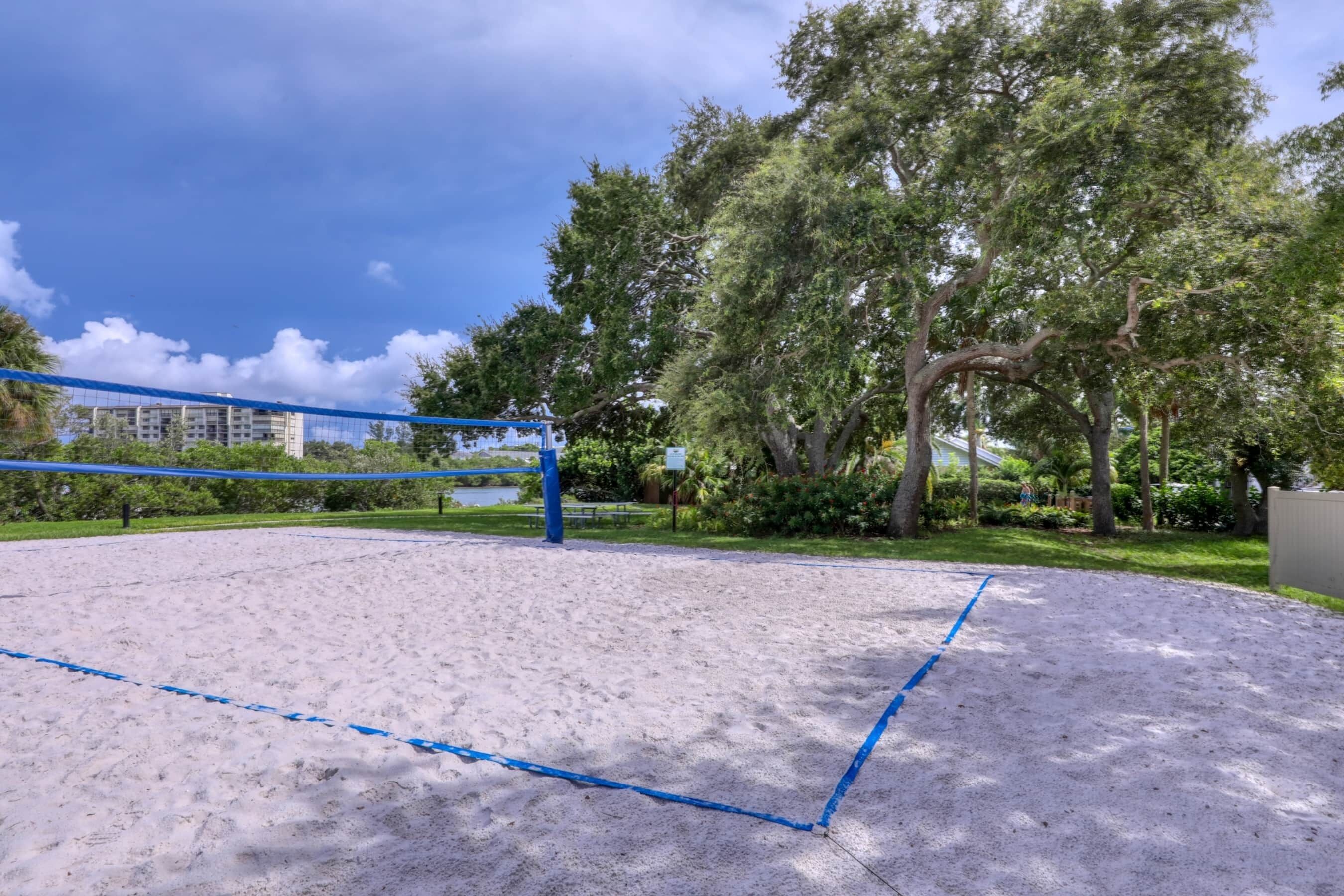 Een zandvolleybalveld met blauwe lijnen, omringd door bomen en een gebouw op de achtergrond.