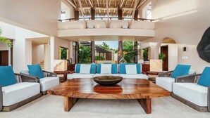 A spacious lounge area with a large wooden coffee table, white sofas with blue cushions, and a view of a tropical garden through large windows.