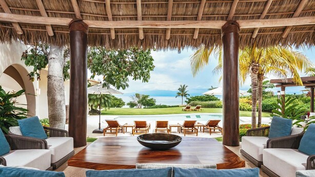 A spacious outdoor seating area with a wooden table and chairs, overlooking a pool and lush greenery.