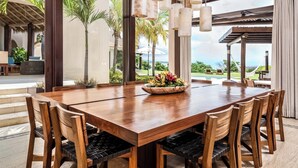 A large wooden dining table with chairs, a flower centerpiece, and a view of a pool and greenery through large windows.