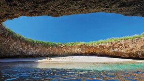 A beach under a rock formation with clear water and a sandy shore.