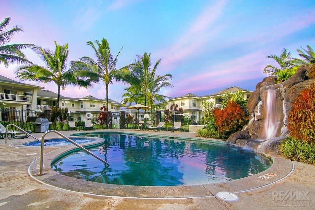 A swimming pool with a waterfall feature, surrounded by palm trees and a building with a balcony.