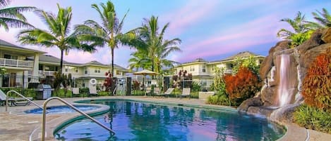 A swimming pool with a waterfall feature, surrounded by palm trees and a building with a balcony.