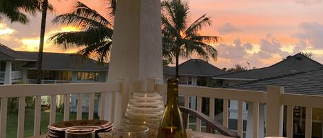 A wooden table with a bottle of wine, a glass of white wine, and a glass of yellow beverage, set against a backdrop of palm trees and houses under a sunset sky.