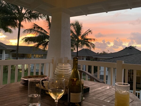 A wooden table with a bottle of wine, a glass of white wine, and a glass of yellow beverage, set against a backdrop of palm trees and houses under a sunset sky.