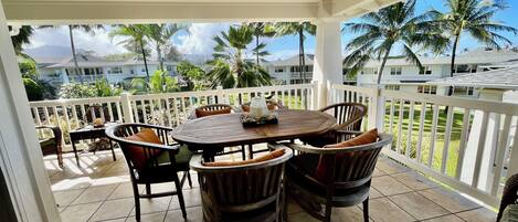 A covered patio with a round wooden table and chairs, a white railing, and a view of palm trees and houses.