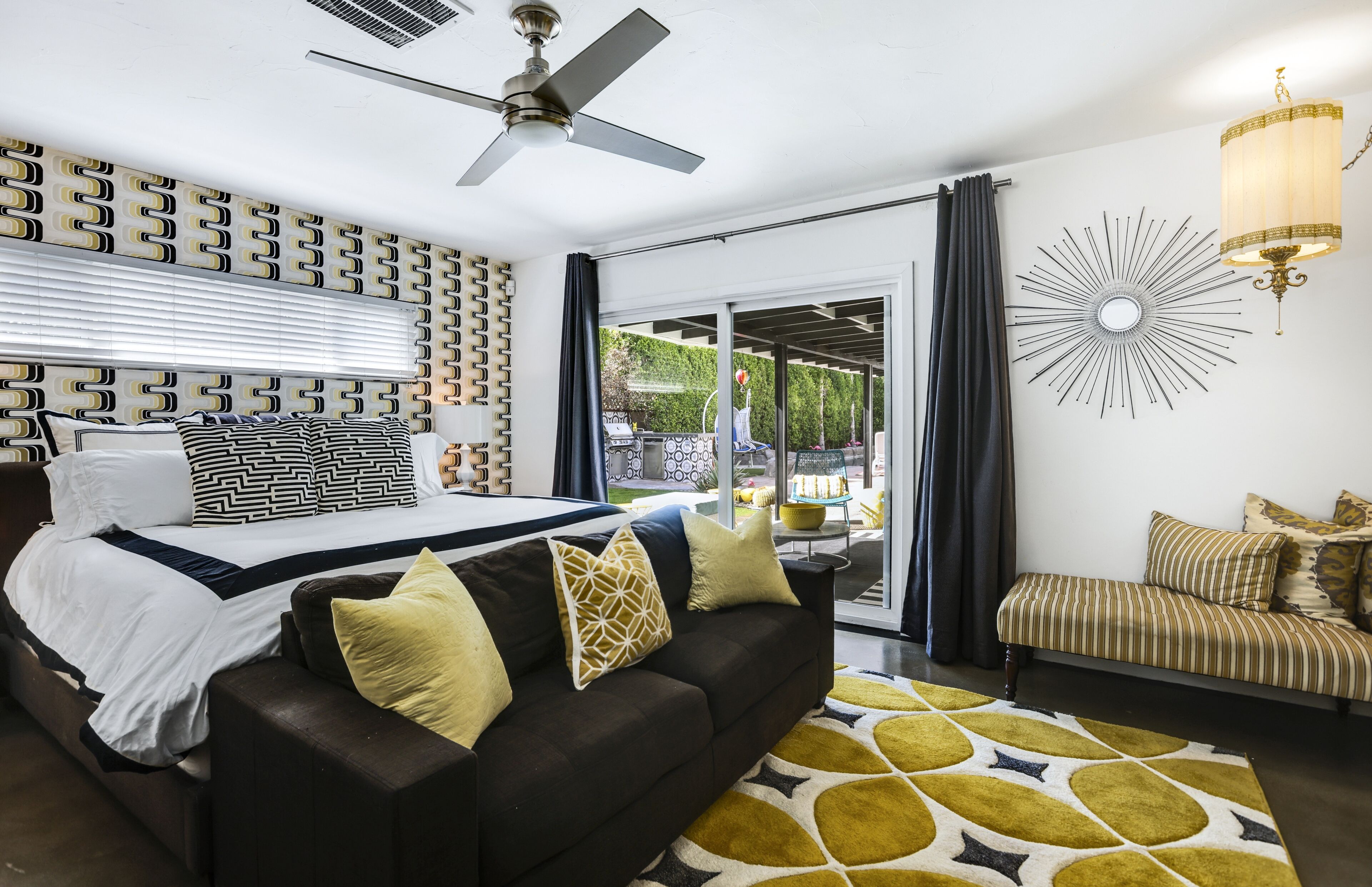 A bedroom with a bed, a sofa, a ceiling fan, a chandelier, and a view through a glass door.