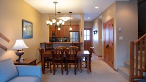 A dining area with a wooden table and chairs, a chandelier, and a kitchen in the background.